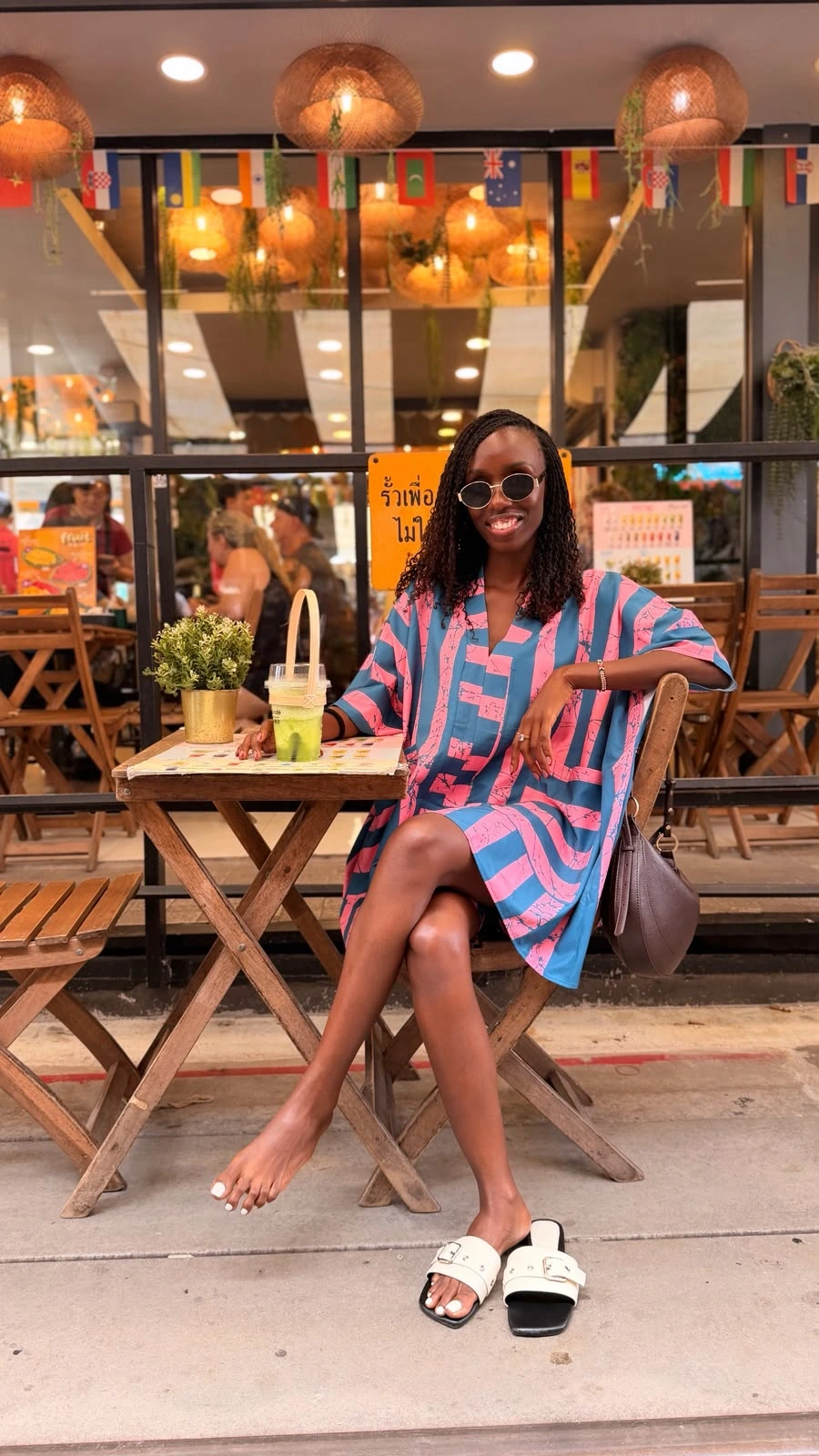 ZuriNiah smiling at an outdoor café in a pink and teal dress, sunglasses, and white sandals