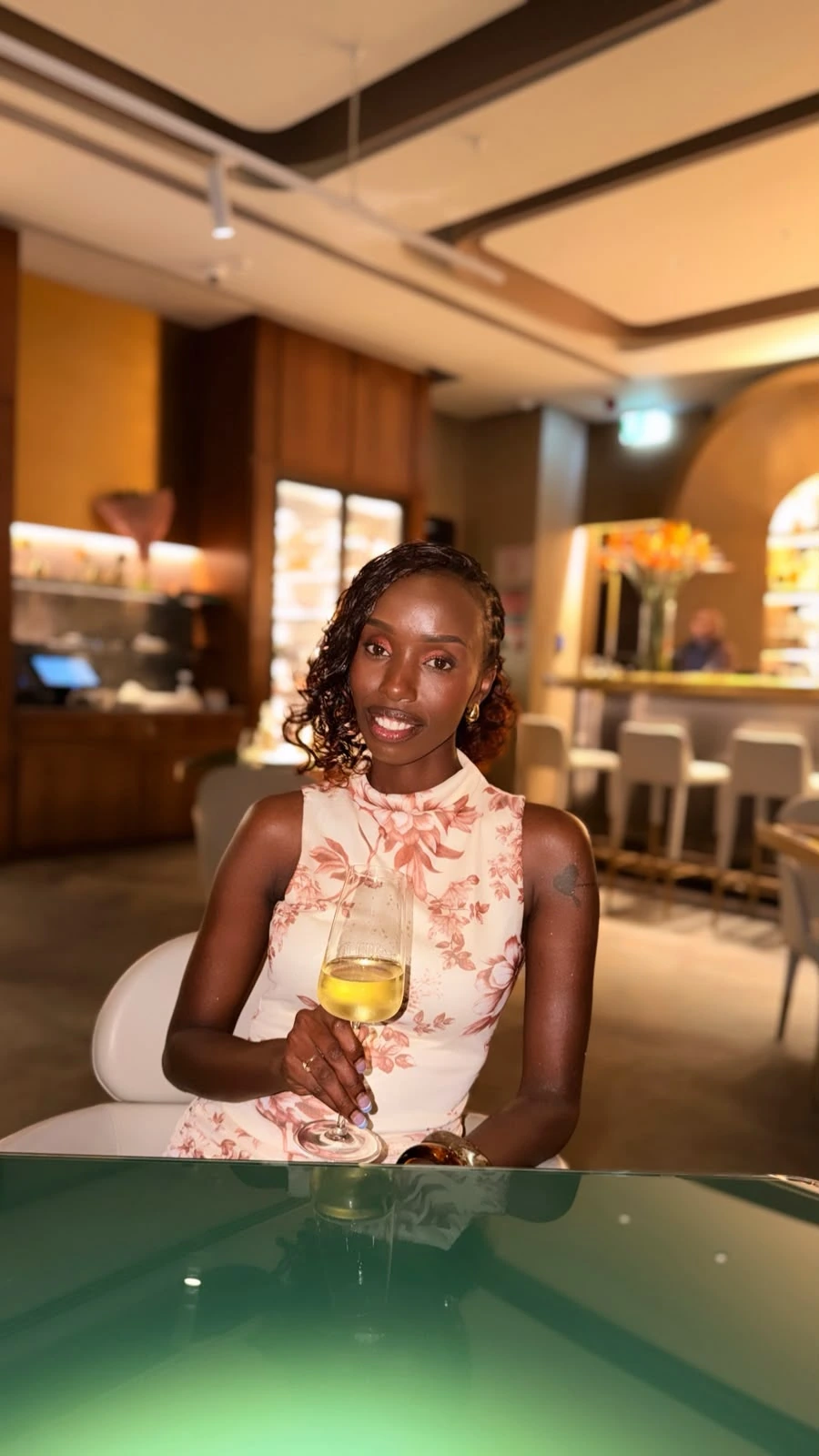 ZuriNiah seated at a glass table in a floral blouse, holding a glass of white wine
