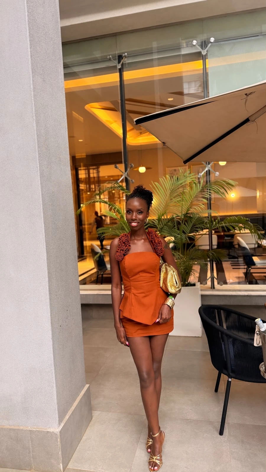 ZuriNiah, Doreen Kathambi Gikundi, in a burnt-orange peplum mini dress and gold heels, holding a gold bag