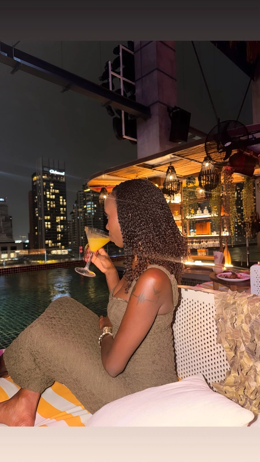 ZuriNiah in profile at a rooftop pool at night, lifting a cocktail, wearing an olive dress