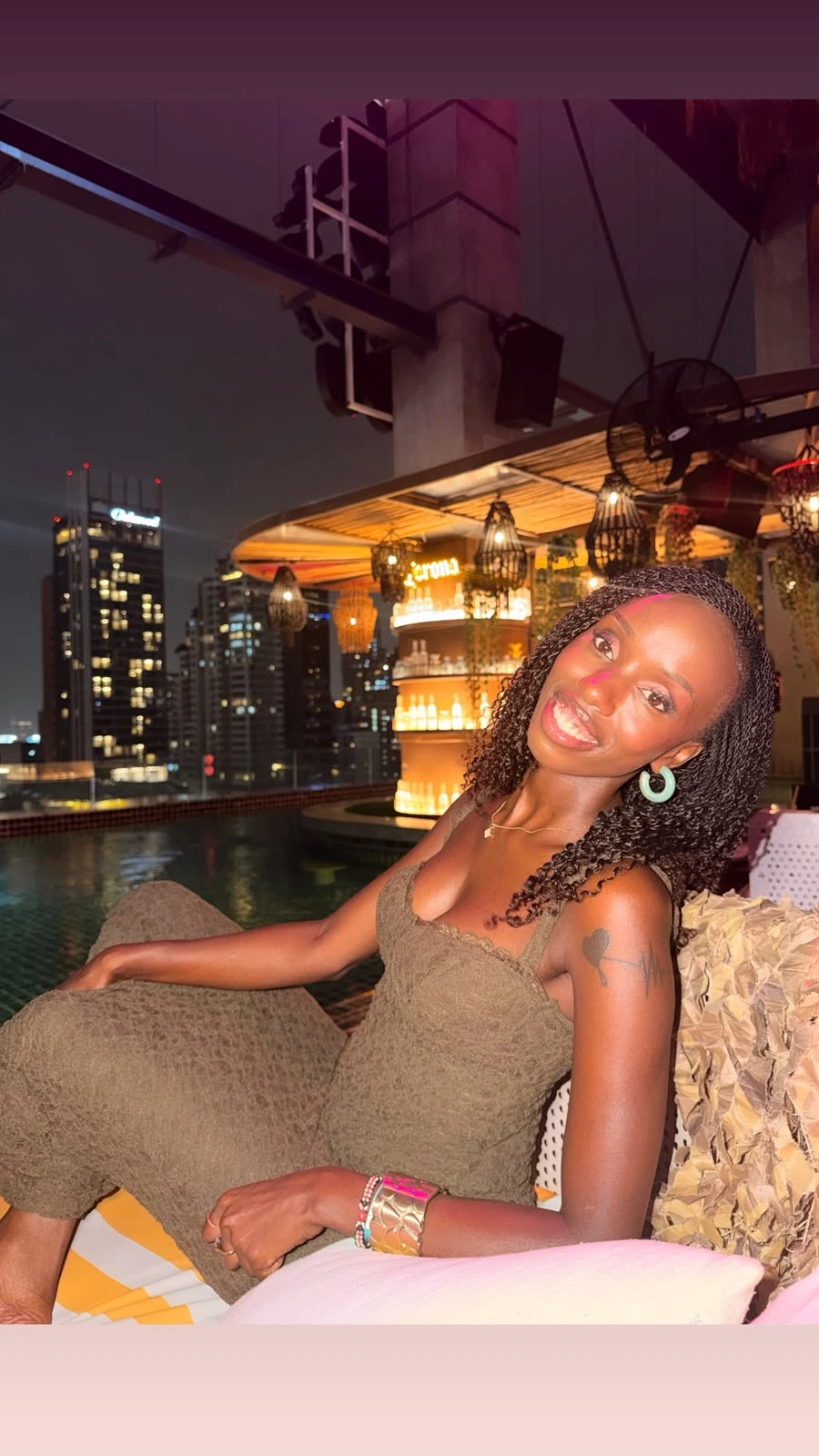 ZuriNiah smiling on a rooftop lounge at night, in an olive dress, with the city skyline behind her