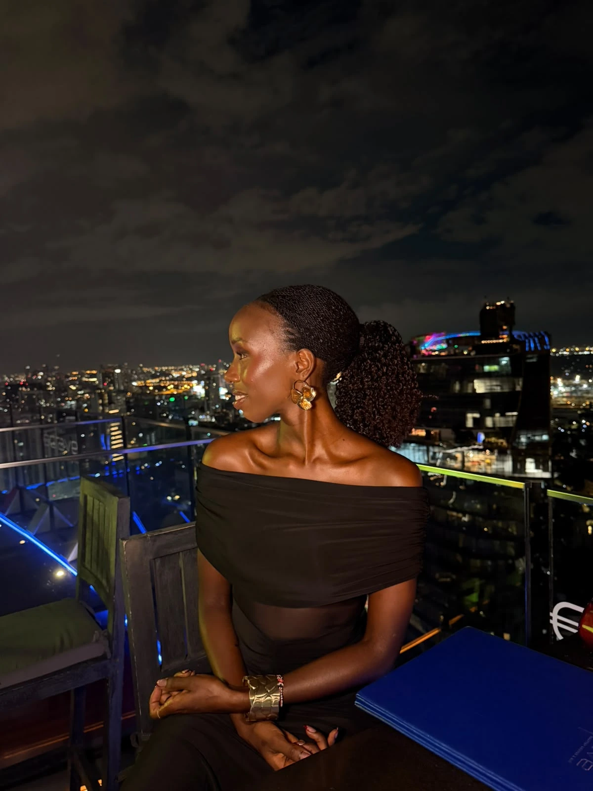 Profile of ZuriNiah on a rooftop at night, hair in a low ponytail, wearing a black off-shoulder dress and gold earrings